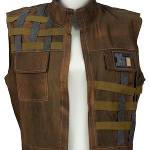 May include: A brown sleeveless vest featuring a high collar, visible stitching, and a woven strap pattern in olive green and dark gray. The garment includes two front pockets, one of which is adorned with a rectangular black and tan badge.