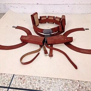 May include: A brown leather utility belt featuring multiple rectangular pouches, a central metallic buckle, and two matching holster attachments with curved leather straps. The set includes additional leather straps with metal buckles and fasteners.