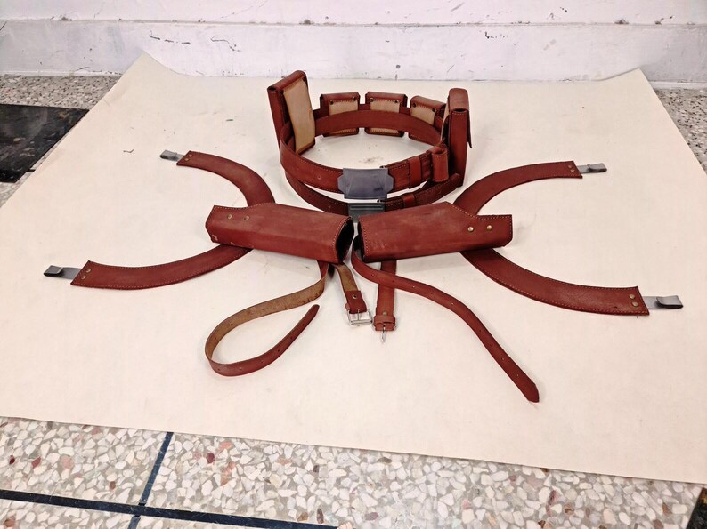 May include: A brown leather utility belt featuring multiple rectangular pouches, a central metallic buckle, and two matching holster attachments with curved leather straps. The set includes additional leather straps with metal buckles and fasteners.