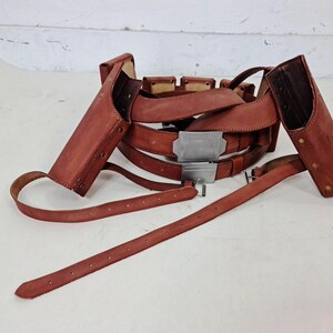 May include: A brown leather gun belt featuring two attached holsters, multiple smaller pouches, and a rectangular silver-toned metal buckle. The belt includes adjustable straps with punched holes and metal hardware, designed to be worn around the waist.