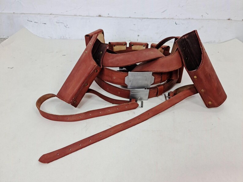 May include: A brown leather gun belt featuring two attached holsters, multiple smaller pouches, and a rectangular silver-toned metal buckle. The belt includes adjustable straps with punched holes and metal hardware, designed to be worn around the waist.