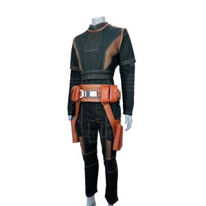 K&ouml;nnte beinhalten: Ein dunkelgr&uuml;nes und braunes Kost&uuml;m mit hohem Kragen, langen &Auml;rmeln und einem braunen G&uuml;rtel mit Taschen. Das Kost&uuml;m umfasst eine passende Hose und zwei braune holsterartige Accessoires.