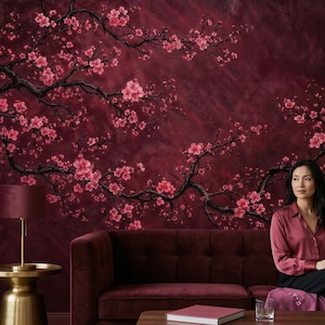 May include: A deep burgundy wall mural depicting cherry blossom branches with pink flowers. The branches spread across the wall, forming a nature-inspired pattern. A burgundy sofa and a woman in a burgundy shirt are in the foreground.