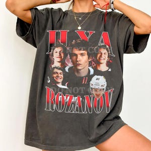 May include: Dark gray t-shirt featuring a graphic of a man with the name "ILYA ROZANOV" in red text. The design includes multiple images of the man, some wearing a hockey helmet. The shirt has a vintage, worn look.
