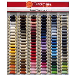 May include: A display of Gütermann Sew-all Thread in various colors. The thread spools are arranged on a rack, with a red and white sign at the top. The sign reads "Premium Quality 100%" and "Gütermann creativ Sew-all Thread 100 m".
