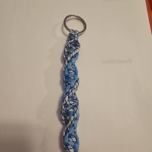 May include: A handmade keychain with a braided design in shades of blue and white. The keychain features a metal ring at the top for attaching keys. The braided section is a series of interwoven knots, creating a textured, decorative accessory.