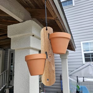 May include: A hanging wooden planter with two terracotta pots. The wooden plank is suspended by a black cord and is attached to a wooden beam. The pots are empty and ready for planting. The planter is hanging outside.