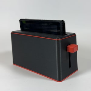 May include: A black and red novelty charging dock shaped like a toaster. The dock has a red trim and a red lever. A black electronic device is inserted into the top slot.