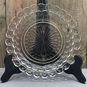 Puede incluir: Un plato de vidrio transparente con un patrón de burbujas alrededor del borde, exhibido en un soporte negro. El plato tiene un borde festoneado y un diseño de estrella en el centro. El fondo es una superficie de madera.