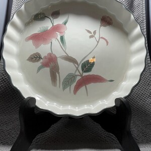 May include: A round, off-white ceramic pie plate with a scalloped edge and a floral design featuring pink and green flowers. The plate is displayed on a black wooden stand.