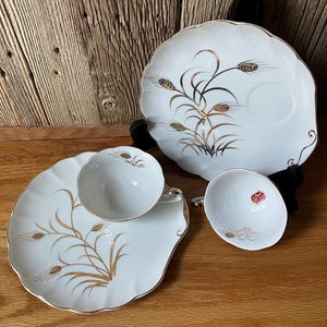 Puede incluir: Dos platillos de porcelana blanca y dos tazas de té a juego con un patrón de tallos de trigo dorados. Los platillos tienen bordes festoneados con ribetes dorados. Una taza de té muestra una pequeña pegatina roja y blanca en el interior.