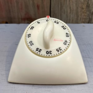 May include: A vintage off-white kitchen timer with a curved handle and a circular dial featuring black numbers from 0 to 55. The Sunbeam brand name is printed in red script on the face, and a small red triangle indicator is at the top center.