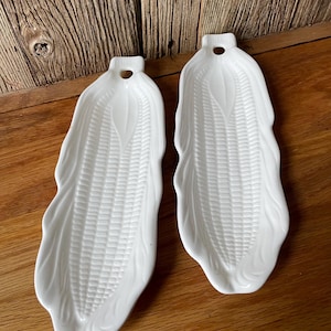 Könnte beinhalten: Zwei weiße Keramik-Servierplatten in Maisform. Jede Platte hat ein detailliertes Design, das die Textur von Maiskörnern und -blättern nachahmt. Die Platten haben oben ein kleines Loch zum Aufhängen. Der Hintergrund ist eine Holzoberfläche.