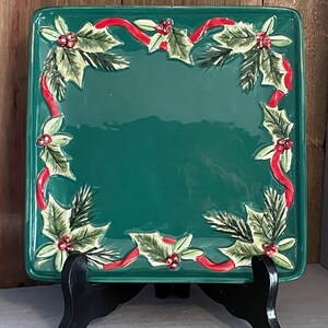 May include: A square, green ceramic serving platter with a festive Christmas design. The border features embossed holly leaves, red berries, and a red ribbon. The platter is displayed on a black stand, ideal for festive occasions.
