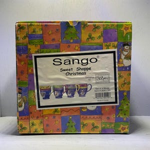 May include: A box of Sango Sweet Shoppe Christmas mugs. The box is decorated with a patchwork pattern of Christmas trees, stars, and snowmen. The box features a label with the Sango logo and the words "Sweet Shoppe Christmas". The set includes four mugs.