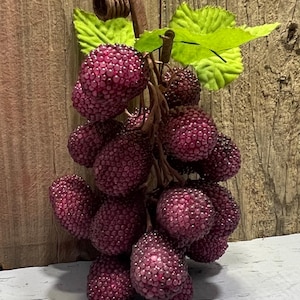 May include: A cluster of artificial grapes with a deep purple hue, featuring a textured surface. The grapes are attached to a brown stem with green leaves. The background is a weathered wooden surface.