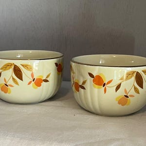 May include: Two cream-colored ceramic bowls with a gold rim and floral design. The floral pattern features orange, yellow, and brown leaves and flowers. The bowls are round and have a ribbed texture.