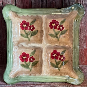 May include: A square ceramic serving dish with four compartments. The dish has a light green border and a beige interior. Each compartment features a red floral design with green leaves. The dish is likely used for serving appetizers or snacks.