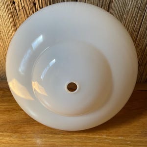 May include: A white, opaque glass light fixture shade. The shade is circular with a rounded, bowl-like shape and a central hole. The surface is smooth and glossy, reflecting light. The fixture is likely for a ceiling light.