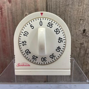 May include: A white Sunbeam kitchen timer with a circular dial featuring black numbers from 0 to 55. The device has a central rotating knob, a red triangular indicator at the top, and a rectangular base. The brand name Sunbeam is printed in red.