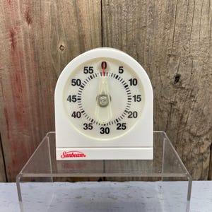 May include: A white Sunbeam kitchen timer with an arched top and a circular face. The dial features black numerals from 0 to 55 in increments of five, with a central clear plastic pointer and a thin red indicator line set at the zero position.
