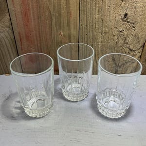 May include: Three clear, vintage-style drinking glasses. Each glass has a textured, vertical ribbed design and a decorative base. The glasses are made of clear glass and are displayed on a light gray surface with a wooden background.