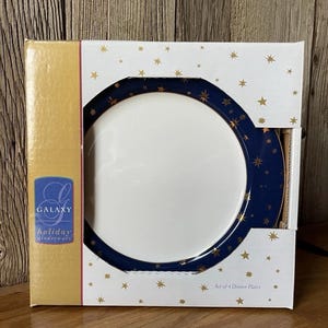 Op de afbeelding: Een set van 4 dinerborden in een witte kartonnen doos met gouden sterpatronen. De borden hebben een donkerblauwe rand met gouden steraccenten en een wit midden. De doos toont de tekst GALAXY holiday dinnerware en Set of 4 Dinner Plates.