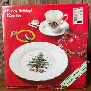 May include: A red cardboard box for a Happy Holidays 12pc. Set featuring a white plate and teacup with saucer, both decorated with a Christmas tree graphic. The box displays a Santa figurine, a yellow wreath, and a red candle with gold tinsel.