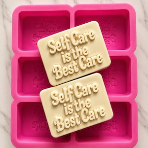 Può includere: Uno stampo in silicone rosa acceso con sei cavità rettangolari, ognuna impressa con il testo "Self Care is the Best Care". Due saponette rettangolari color bianco sporco con lo stesso testo in rilievo sono posizionate sopra lo stampo.