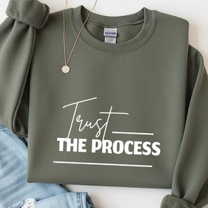 Op de afbeelding: Een olijfgroene sweatshirt met de woorden "Trust the Process" in witte letters. Een goudkleurige ketting met een ronde hanger ligt op de sweatshirt. De sweatshirt is op een blauwe spijkerbroek gevouwen.