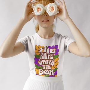 May include: White t-shirt with the text "THE KNIFE STAYS IN THE BOX" in a retro font, surrounded by orange, purple, and yellow flowers. The person is wearing a white skirt and holding two white roses in front of their eyes.