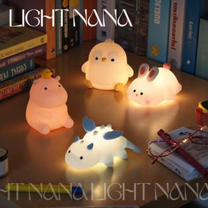 Può includere: Una collezione di luci notturne luminose in stile cartone animato a forma di ippopotamo, pinguino, coniglietto e dinosauro. Le luci sono soffuse ed emettono un bagliore caldo, perfetto per la cameretta di un bambino. Il testo "LIGHT NANA" è in alto.