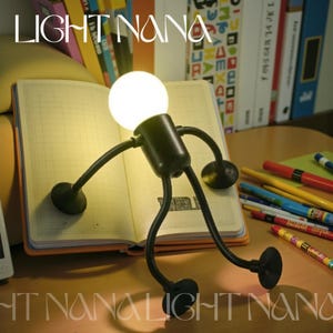 May include: A quirky black desk lamp in the form of a stick figure, featuring a spherical light bulb as a head. The lamp is placed on an open notebook. The text "LIGHT NANA" is visible. Colourful pens and books are in the background.