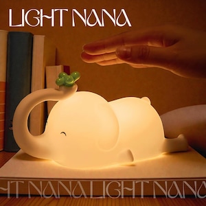 May include: A soft, glowing white elephant-shaped night light with a small green accent on its trunk. The lamp is on a wooden surface, with the text "LIGHT NANA" above and "HT NANALIGHT NANA" below. A hand hovers above the lamp.