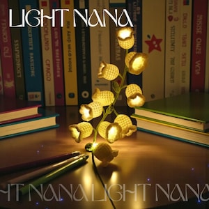 May include: A string of warm white LED fairy lights shaped like bell flowers. The lights are on a green wire, with the text "LIGHT NANA" in the top left and bottom right corners. The lights are on a wooden surface with books in the background.