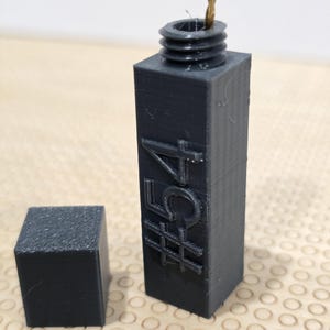 May include: A dark gray, rectangular container with a threaded top and a separate cube. The container has the number "#54" embossed on its side. A small, thin, brown object protrudes from the top.