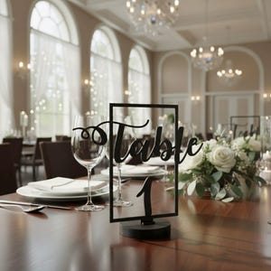 May include: A black table number sign with the word "Table" in cursive script and the number "1". The sign is in a rectangular frame and sits on a round base. The table is set with plates, silverware, and wine glasses.