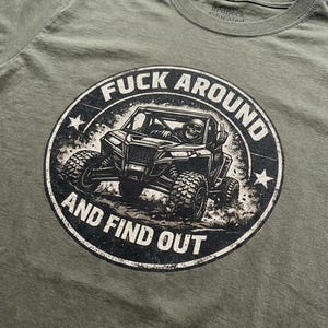 May include: Olive green t-shirt with a distressed graphic. The graphic is a circle with the text "FUCK AROUND AND FIND OUT" around an image of an off-road vehicle with a skeleton driver giving the middle finger.