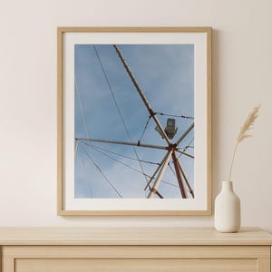 May include: A framed photograph of a Ferris wheel structure against a blue sky. The metal framework is red and white, with a spotlight and cables. The photograph is in a light wood frame, and a vase with a plant is on a wooden surface.