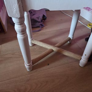 Puede incluir: Primer plano de una silla de madera pintada de blanco con cuatro patas. Las patas están conectadas por dos travesaños de madera natural. La silla está sobre un suelo de madera marrón claro.