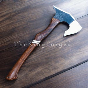 Könnte beinhalten: Eine handgefertigte Axt mit einem dunklen Holzgriff und einer silberfarbenen Klinge. Der Griff hat weiße Akzente und kleine dekorative Nieten. Der Text "The King Forged" ist auf dem Griff aufgedruckt.