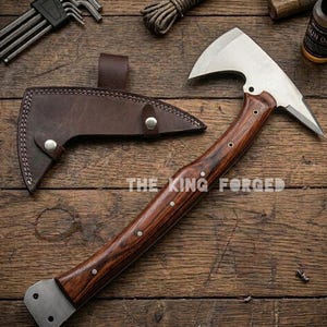 Puede incluir: Un hacha artesanal con mango de madera y hoja de acero inoxidable. El hacha tiene una funda de cuero marr&oacute;n y el texto "THE KING FORGED" es visible. El hacha se muestra sobre una superficie de madera.