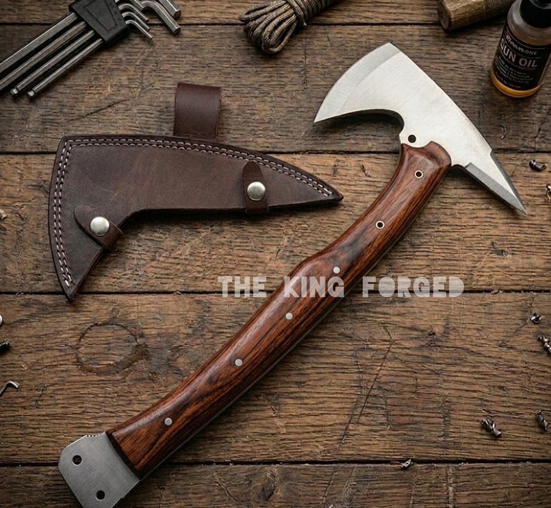 Puede incluir: Un hacha artesanal con mango de madera y hoja de acero inoxidable. El hacha tiene una funda de cuero marr&oacute;n y el texto "THE KING FORGED" es visible. El hacha se muestra sobre una superficie de madera.