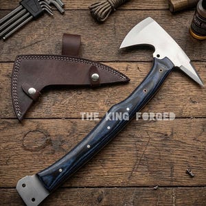 Puede incluir: Un hacha forjada con una hoja plateada y un mango de madera azul oscuro. El hacha tiene una funda de cuero y el texto "THE KING FORGED" es visible. El hacha est&aacute; sobre una superficie de madera.
