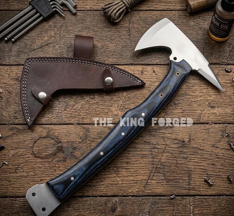Puede incluir: Un hacha forjada con una hoja plateada y un mango de madera azul oscuro. El hacha tiene una funda de cuero y el texto "THE KING FORGED" es visible. El hacha est&aacute; sobre una superficie de madera.