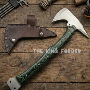 Puede incluir: Un hacha de acero forjado con un mango de madera verde y negro y una funda de cuero marr&oacute;n. La cabeza del hacha es plateada y tiene una hoja afilada. El texto "THE KING FORGED" est&aacute; impreso en la madera.