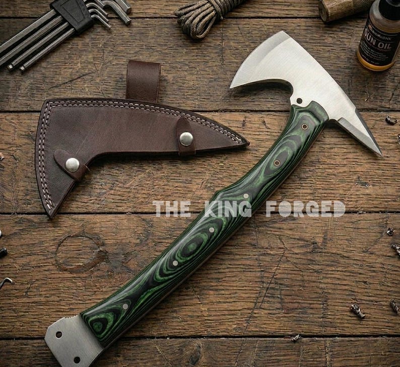 Puede incluir: Un hacha de acero forjado con un mango de madera verde y negro y una funda de cuero marr&oacute;n. La cabeza del hacha es plateada y tiene una hoja afilada. El texto "THE KING FORGED" est&aacute; impreso en la madera.