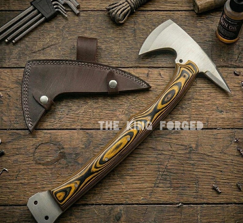 Puede incluir: Un hacha t&aacute;ctica con una hoja de acero inoxidable y un mango de madera con vetas negras y amarillas. Incluye una funda de cuero marr&oacute;n. El texto "THE KING FORGED" es visible.