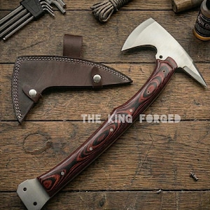 Puede incluir: Un hacha t&aacute;ctica con una hoja de acero inoxidable y un mango de madera con vetas rojas y negras. El hacha va acompa&ntilde;ada de una funda de cuero marr&oacute;n. El texto "THE KING FORGED" es visible en el mango.