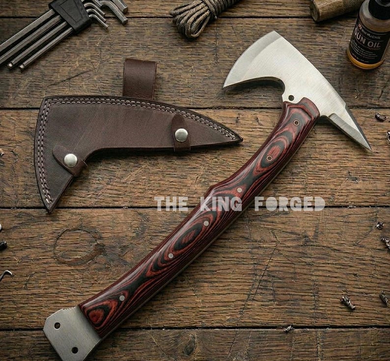 Puede incluir: Un hacha t&aacute;ctica con una hoja de acero inoxidable y un mango de madera con vetas rojas y negras. El hacha va acompa&ntilde;ada de una funda de cuero marr&oacute;n. El texto "THE KING FORGED" es visible en el mango.